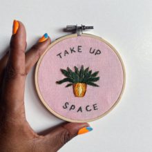 a dark-skinned hand with colorful nail polish holing an embroidery composed of text that reads “Take up space” and a potted plant design accenting the text