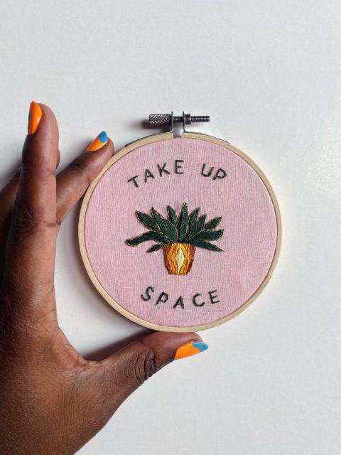 a dark-skinned hand with colorful nail polish holing an embroidery composed of text that reads “Take up space” and a potted plant design accenting the text