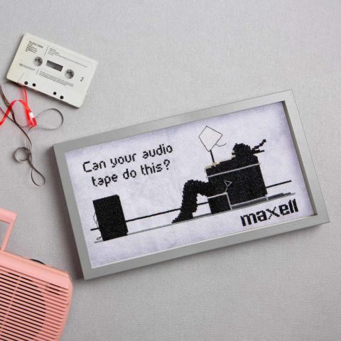 cross-stitch of an old Maxell casette tape ad showing a man's hair being blown back by a speaker and the words "can your audio tape do this?"