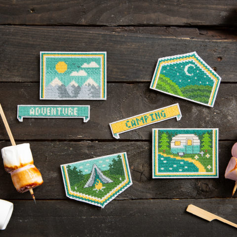 A set of small cross-stitched camping-themed badges on a wooden table next to roasted marshmallows