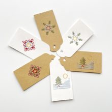 Three brown kraft paper and three white paper gift tags colorfully embroidered with images of two snowflakes and a tree on a snowy mountain