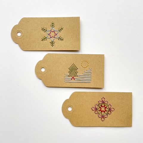 Three brown kraft paper gift tags colorfully embroidered with images of two snowflakes and a tree on a snowy mountain