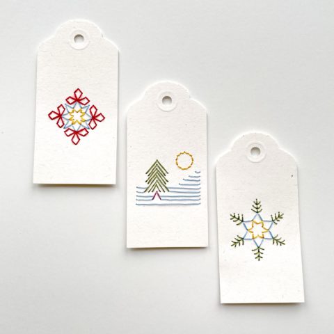 Three white paper gift tags colorfully embroidered with images of two snowflakes and a tree on a snowy mountain