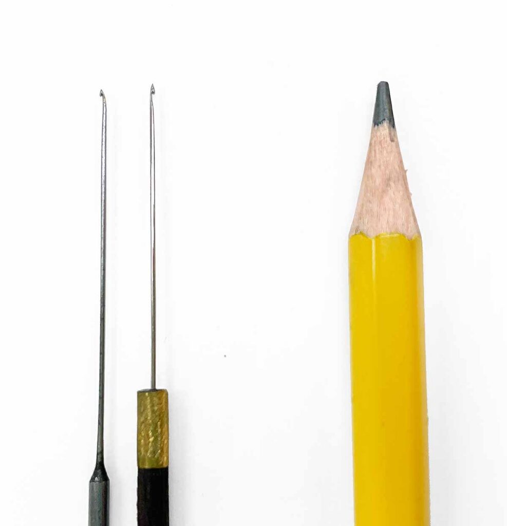 two very fine hooked aari needles shown next to a pencil tip