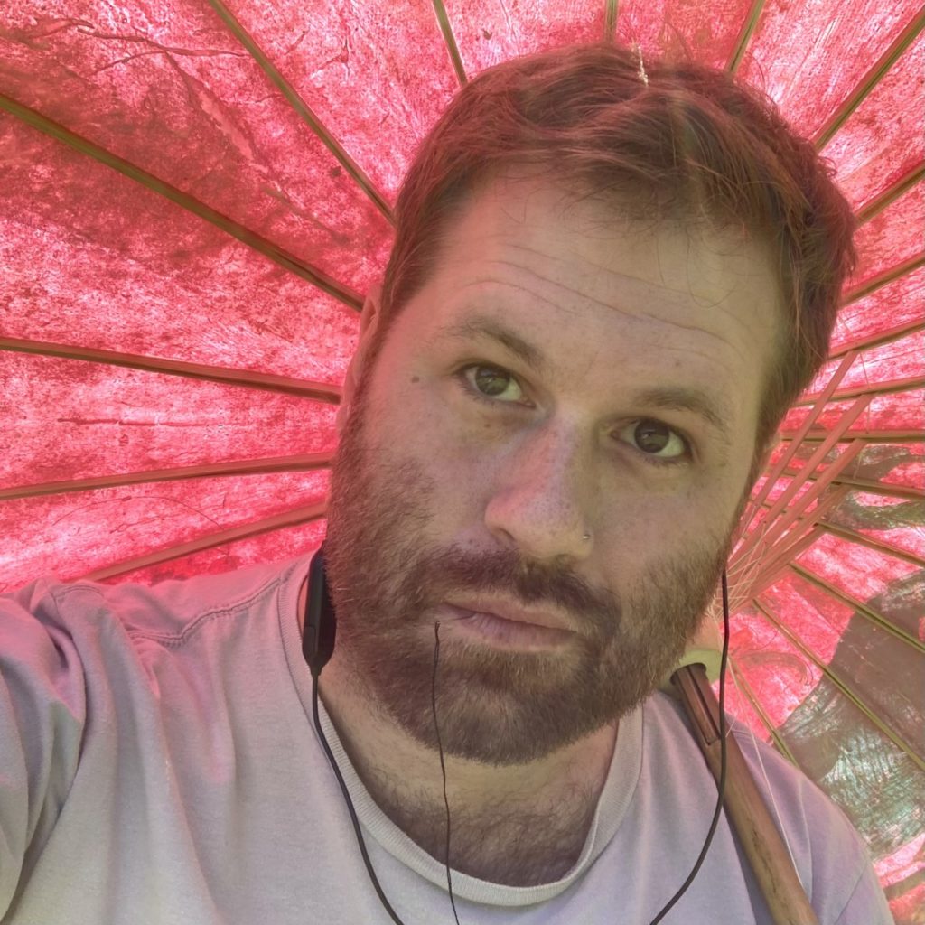artist tom katsumi's face shown under a pink paper umbrella, wearing headphones and with a threaded needle dangling from his mouth