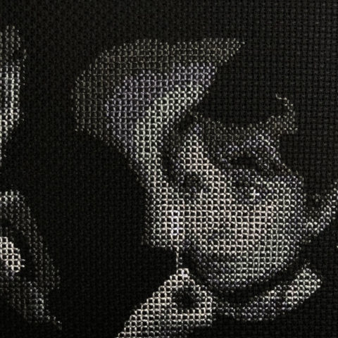 A closeup of audrey hepburn's face illuminated by a man holding up a match stitched in grayscale on black fabric