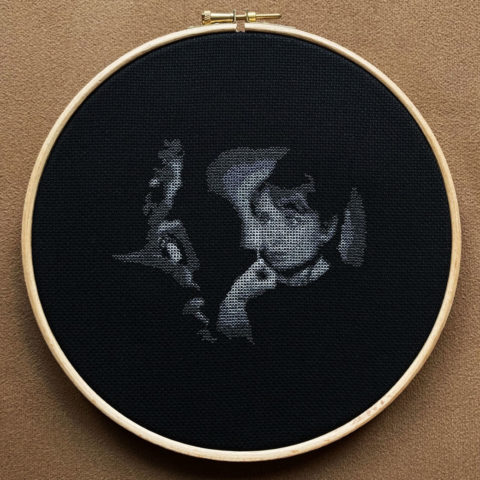 audrey hepburn's face illuminated by a man holding up a match stitched in grayscale on black fabric in a wooden embroidery hoop