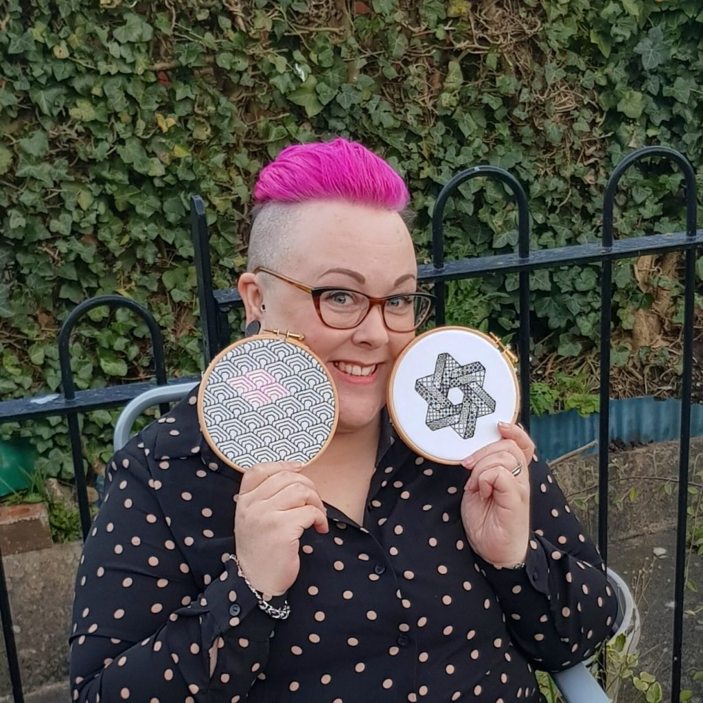 Artist Claire Laurie holding two examples of her needle art in front of a leafy background