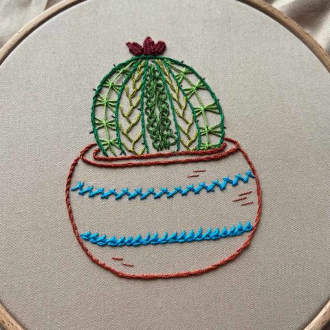 embroidery of a cactus in a striped terra cotta pot stitched on grayish tan fabric inside a wooden embroidery hoop