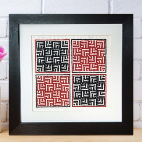 A cross stitch design in red and black on white showing a basketweave grid of nested right angles and displayed in a black wood frame