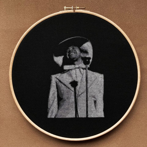 cab calloway spotlit behind a microphone stitched in grayscale on black fabric in a wooden embroidery hoop