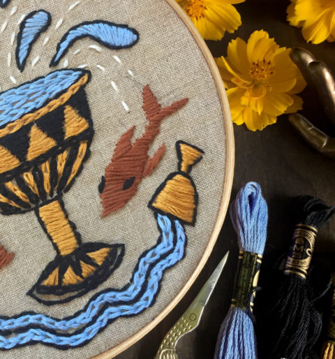 close-up of tan fabric in a wooden embroidery hoop stitched with blue and brown designs of overflowing cups and two koi fish