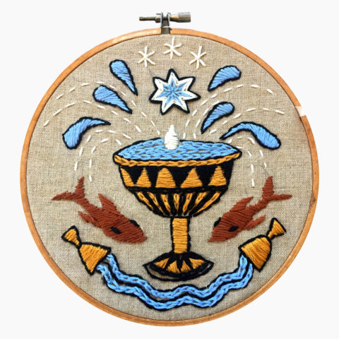tan fabric in a wooden embroidery hoop stitched with blue and brown designs of overflowing cups and two koi fish