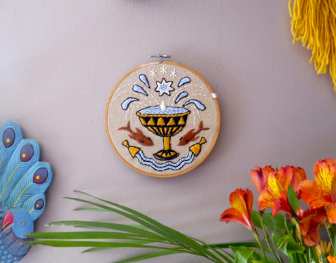 tan fabric in a wooden embroidery hoop stitched with blue and brown designs of overflowing cups and two koi fish mounted on a grey wall next to flowers and corners of other decor pieces