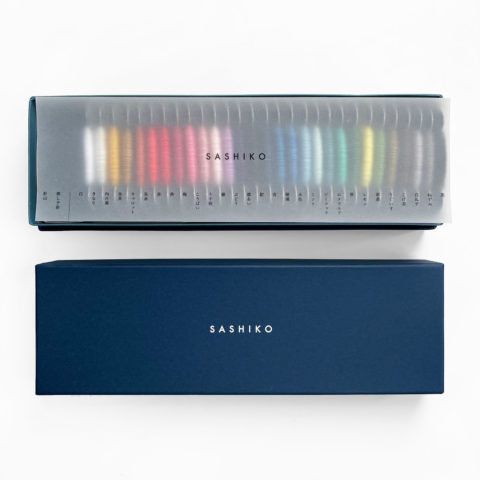 Sashiko thread bobbins in 29 colors arranged in a rainbow inside the bottom of a blue paper gift box. Laid on top is a sheet of vellum with the word "sashiko" and a contents list in Japanese. Displayed below is the lid of the box, which has the word "sashiko" stamped in white.