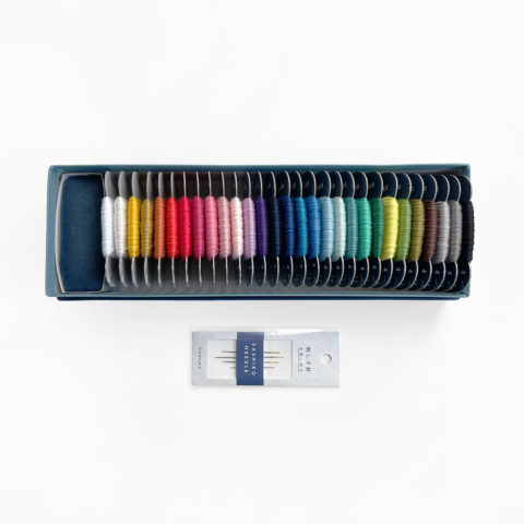 Sashiko thread bobbins in 29 colors arranged in a rainbow and a small dark blue pincushion inside the bottom of a blue paper gift box. Displayed below is a package of 4 assorted sashiko needles