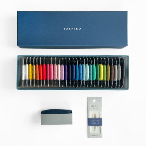 Sashiko thread bobbins in 29 colors arranged in a rainbow inside the bottom of a blue paper gift box. Displayed above is the lid of the box, which has the word "sashiko" stamped in white. Displayed below is a dark blue pincushion inside grey paper and package of 4 assorted sashiko needles