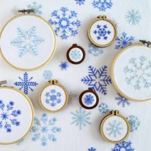 Snowflakes of various types and sizes embroidered in blue on white fabric, some displayed in small wooden hoops