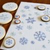 Snowflakes of various types and sizes embroidered in blue on white fabric, some displayed in small wooden hoops on a wooden table