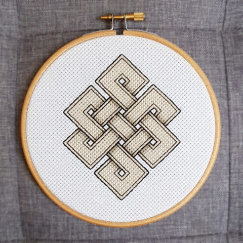 eternal knot pattern cross-stitched in beige and black on white fabric. Displayed in a blond wood embroidery hoop on a grey fabric background