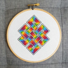 eternal knot pattern cross-stitched in rainbow colors on white fabric. Displayed in a blond wood embroidery hoop on a grey fabric background