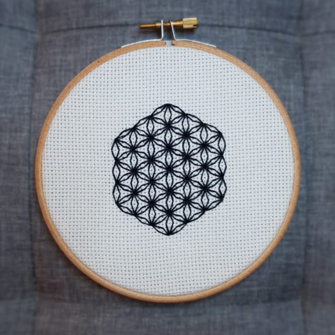 flower of life mystical circle design stitched using blackwork embroidery on white aida canvas. Displayed in a blond wood embroidery hoop on a grey background