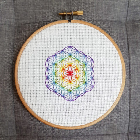 flower of life mystical circle design stitched using blackwork embroidery in rainbow colors on white aida canvas. Displayed in a blond wood embroidery hoop on a grey background