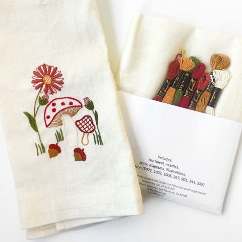 mushrooms, flowers, and acorns embroidered on an ivory linen tea towel displayed next to the fabric and threads needed to make it