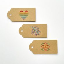 Three brown kraft paper gift tags colorfully embroidered with images of a rainbow heart, a branch, and a poinsettia