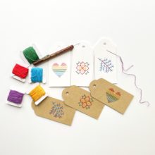 Three brown kraft paper and three white paper gift tags colorfully embroidered with images of a rainbow heart, a branch, and a poinsettia, with stitching supplies nearby