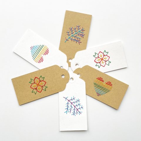 Three brown kraft paper and three white paper gift tags colorfully embroidered with images of a rainbow heart, a branch, and a poinsettia