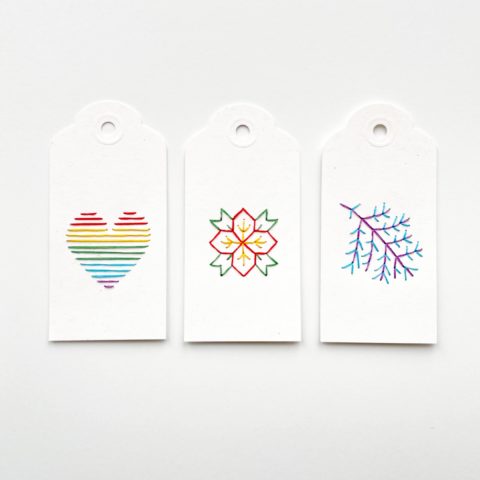 Three white paper gift tags colorfully embroidered with images of a rainbow heart, a branch, and a poinsettia