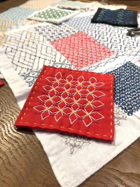 A red fabric coaster stitched with the ajisai-sashi sashiko pattern in red, white, and yellow. Underneath it is a white kitchen cloth with other sashiko patterns stitched in different colors.