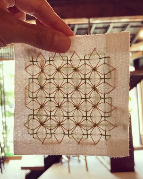 Two fingers holding a white cotton coaster stitched with the ajisa-sashi hydrangea sashiko pattern in blue and cream. The coaster is held up to a sunny window so you can see both the stitches in the front and the back.