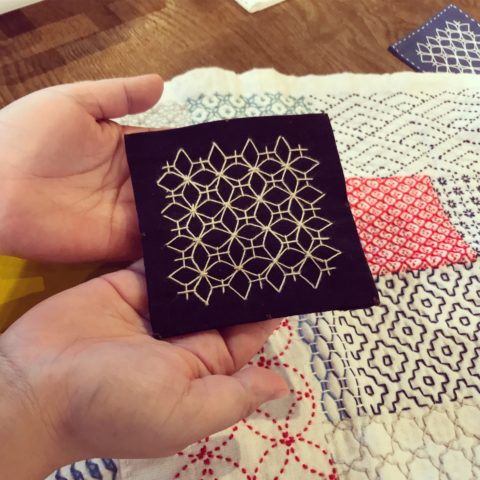 Two hands holding a navy blue fabric coaster stitched with the ajisai-sashi sashiko pattern in white. In the background is a white kitchen cloth with other sashiko patterns stitched in different colors.