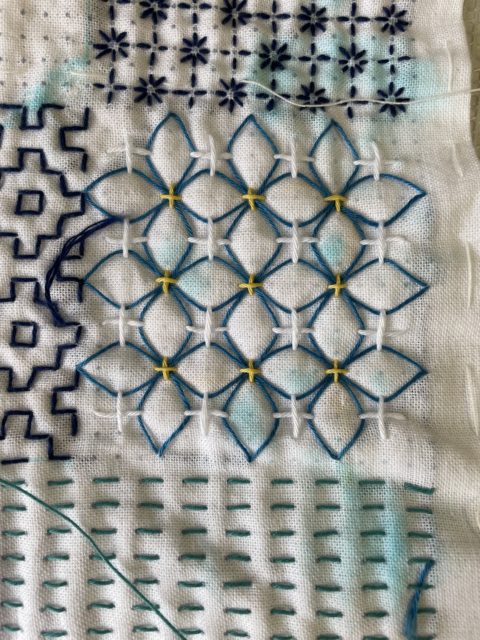 A close-up of ajisai-sashi hydrangea sashiko stitches done in dark blue, white, and yellow on white fabric. You can see bits of other sashiko patterns peeking in at the edges.