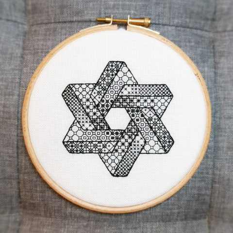 A hexagram made from two intersecting Penrose triangles stitched in blackwork patterns on white canvas. Displayed in a blond wood hoop against a grey background