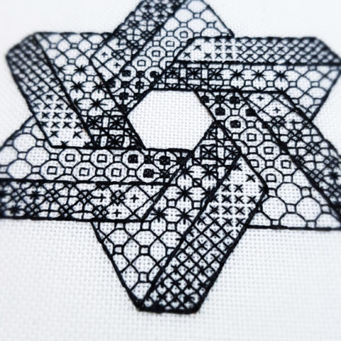 Close-up of the corner of a hexagram made from two intersecting Penrose triangles stitched in blackwork patterns on white canvas.