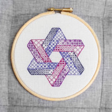 A hexagram made from two intersecting Penrose triangles stitched in purple blackwork patterns on white canvas. Displayed in a blond wood hoop against a grey background