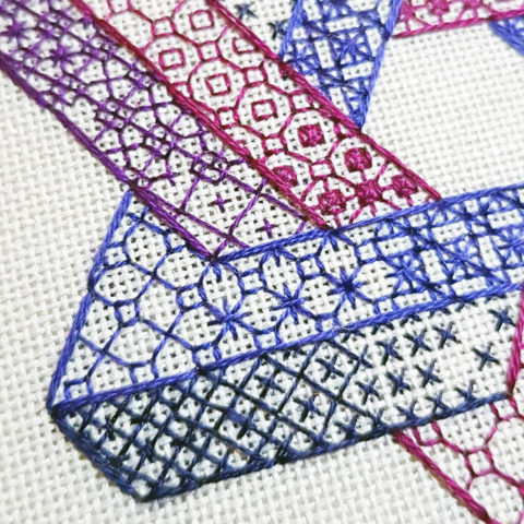 Close-up of the corner of a hexagram made from two intersecting Penrose triangles stitched in purple blackwork patterns on white canvas.