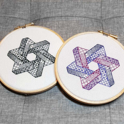 Two hexagrams made from two intersecting Penrose triangles stitched in purple and black blackwork patterns on white canvas. Displayed in blond wood hoops against a grey background