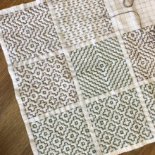 A white square of fabric divided into a 3x3 grid with each square featuring a different variation of the kakinohana persimmon flower sashiko pattern embroidered in green