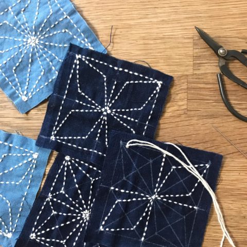 blue fabric squares stitched with the kaku-asanoha hemp flower sashiko pattern scattered on a wooden tabletop next to black scissors
