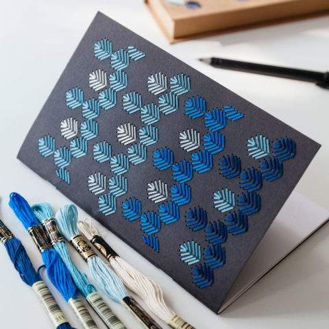A black notebook embroidered with a geometric leaf dsign in various shades of blue shown on a desk beside embroidery floss and a black pencil