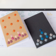 A black and a kraft brown notebook embroidered with a geometric leaf pattern stitched in blues and autumn jewel tones on top of a desk calendar showing the month of July