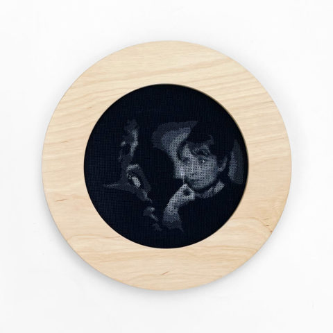 A circular blond wood frame containing a grayscale blackwork image of a film still featuring audrey hepburn staring at a lit match from the movie charade