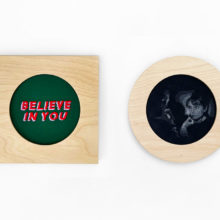 one square and one circular birch wood frame displayed against a white wall. The square frame on the left contains the words "believe in you" embroidered in red and purple on a dark green background. The circular frame on the right contains a grayscale blackwork image of a film still featuring audrey hepburn staring at a lit match from the movie charade
