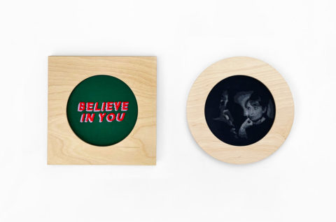 one square and one circular birch wood frame displayed against a white wall. The square frame on the left contains the words "believe in you" embroidered in red and purple on a dark green background. The circular frame on the right contains a grayscale blackwork image of a film still featuring audrey hepburn staring at a lit match from the movie charade