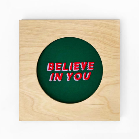 A square, blond wood frame containing the words "believe in you" embroidered in red and purple on a dark green background.