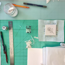 A flower drawn on white fabric stretched around a square block with straight pins seen on a green grid mat surrounded by trimming supplies